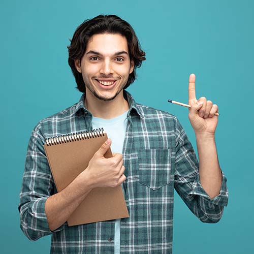 smiling young handsome man holding note pad and pencil looking at camera pointing up isolated on blue background